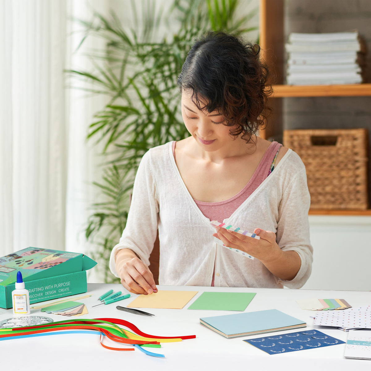 Woman sitting at a table with craft materials, surrounded by plants and books.