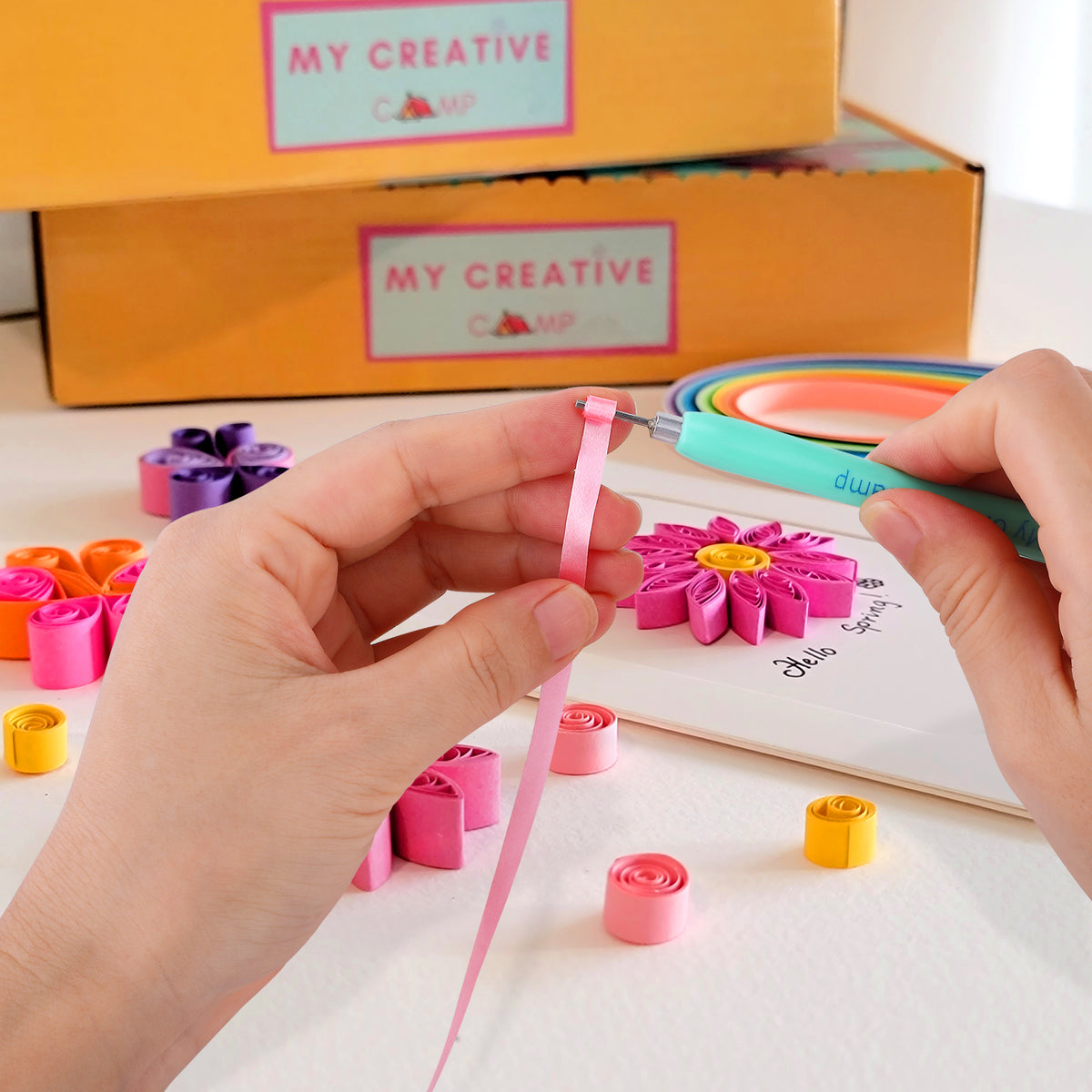 Hands crafting paper flowers with tools and materials on a table, featuring 'My Creative Camp' branding.  paper quilling slotted tool.