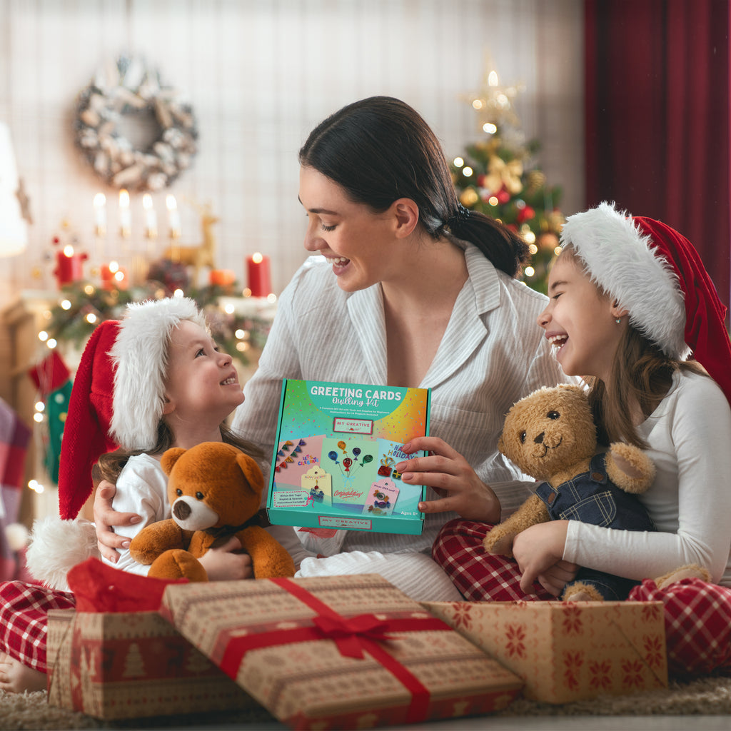 Woman and two children with Christmas hats, surrounded by presents and a box of greeting cards, in a festive setting.
Parent and children making handmade greeting cards using a beginner paper quilling craft kit